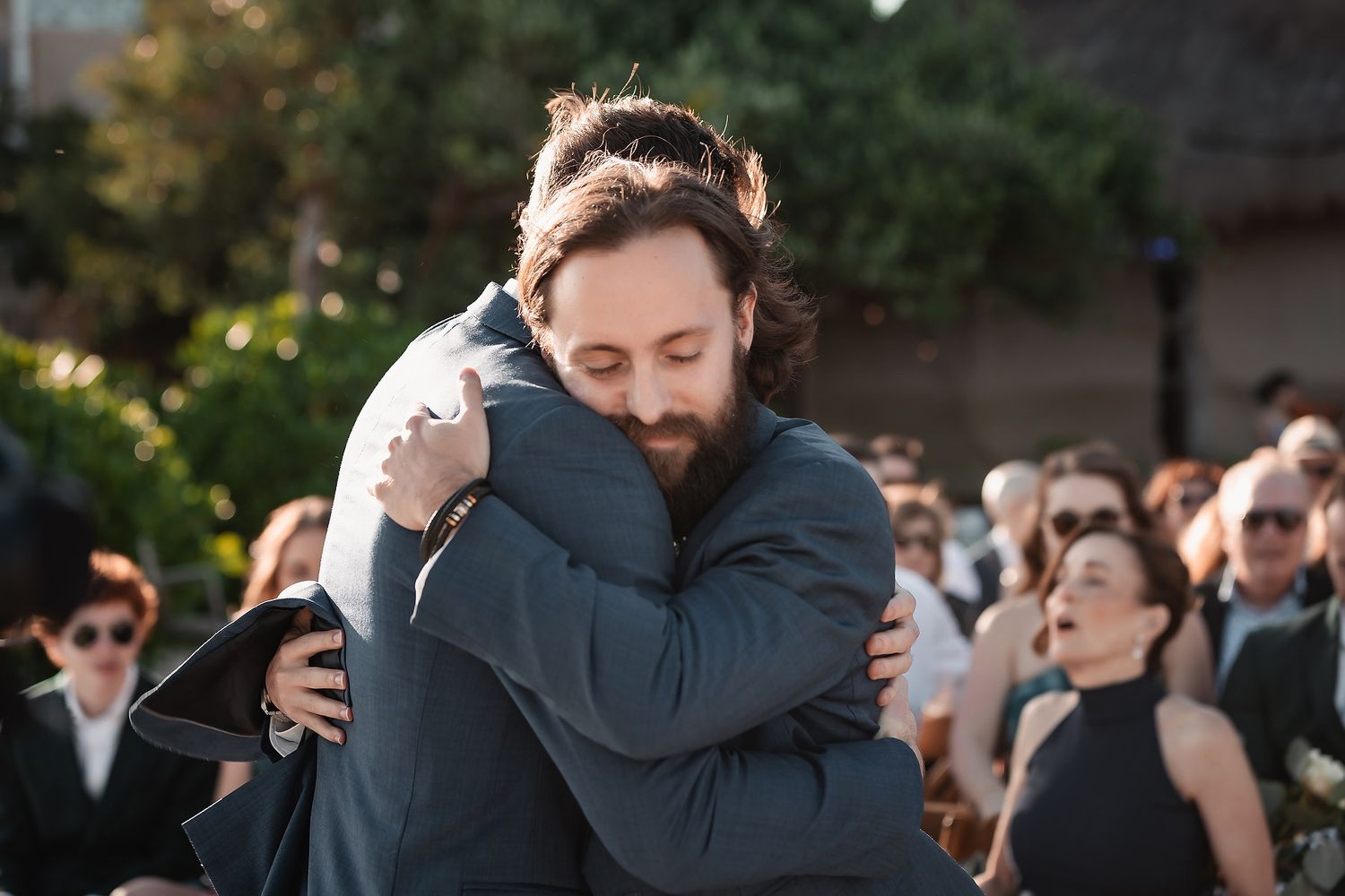 Emotional moment as bearded groom hugs guest during outdoor wedding ceremony with crowd behind.