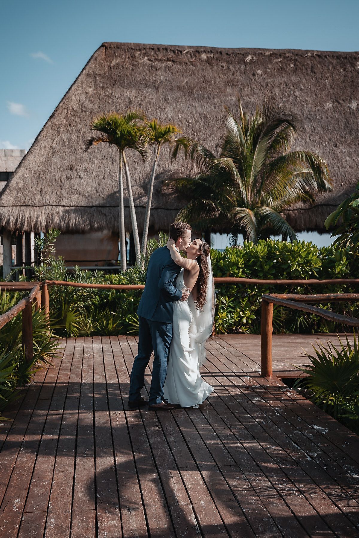 Bride and groom share a romantic kiss on a wooden deck surrounded by tropical palms and thatched roof at a destination wedding.