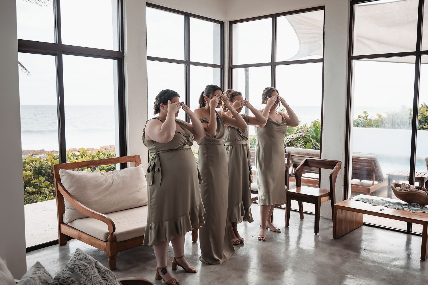 Four bridesmaids in olive dresses adjusting their hair in a bright room with ocean views.