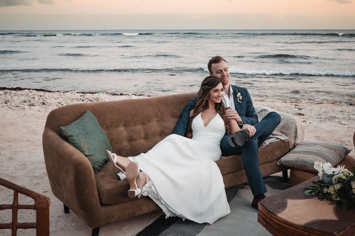 Bride and groom relax on vintage sofa on beach at sunset during wedding reception.