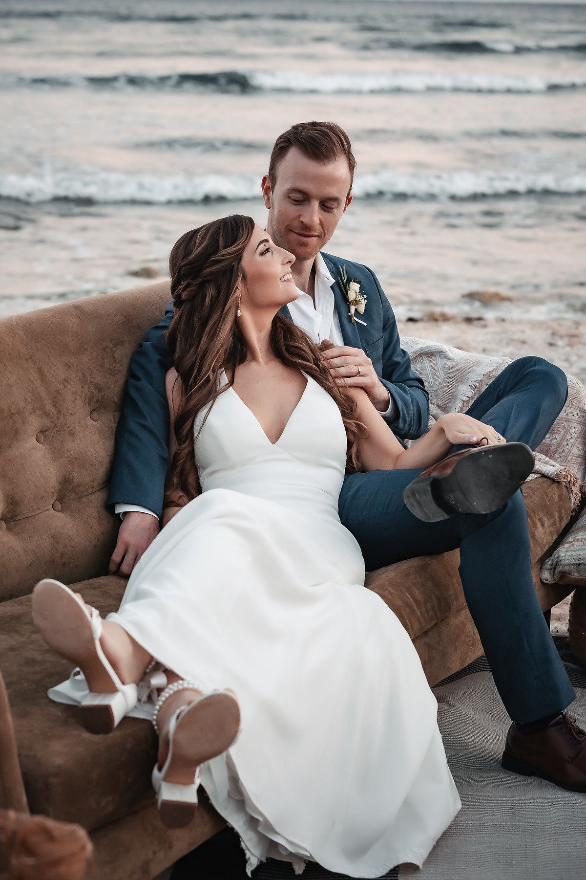 Bride and groom sitting together on a vintage sofa on the beach, sharing a romantic moment in wedding attire.