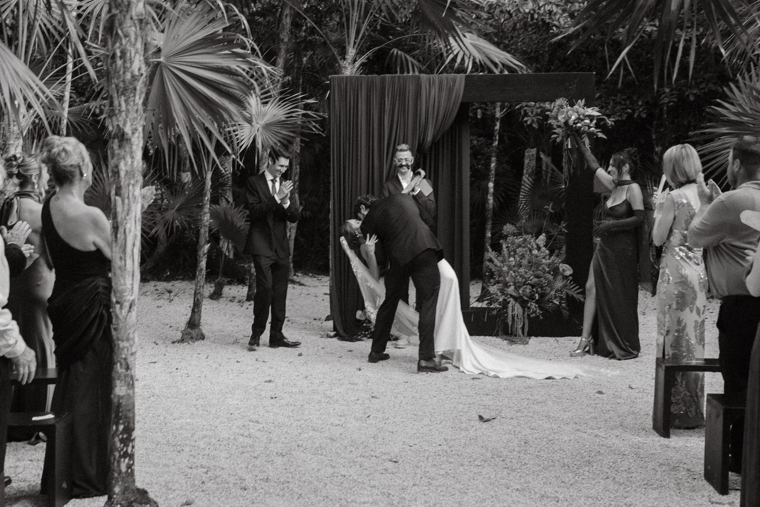 Black and white outdoor wedding ceremony with bride, groom, and guests near tropical palms and dark draped altar.