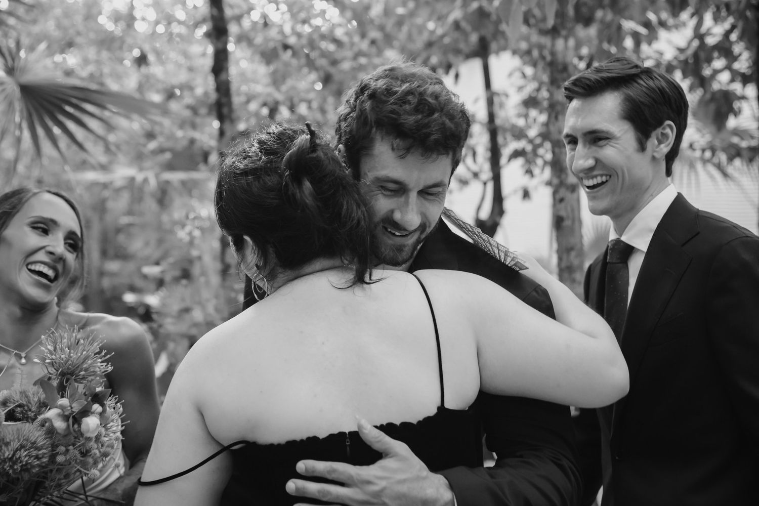 Black and white wedding photo of groom hugging guest outdoors, with smiling bridal party members nearby.