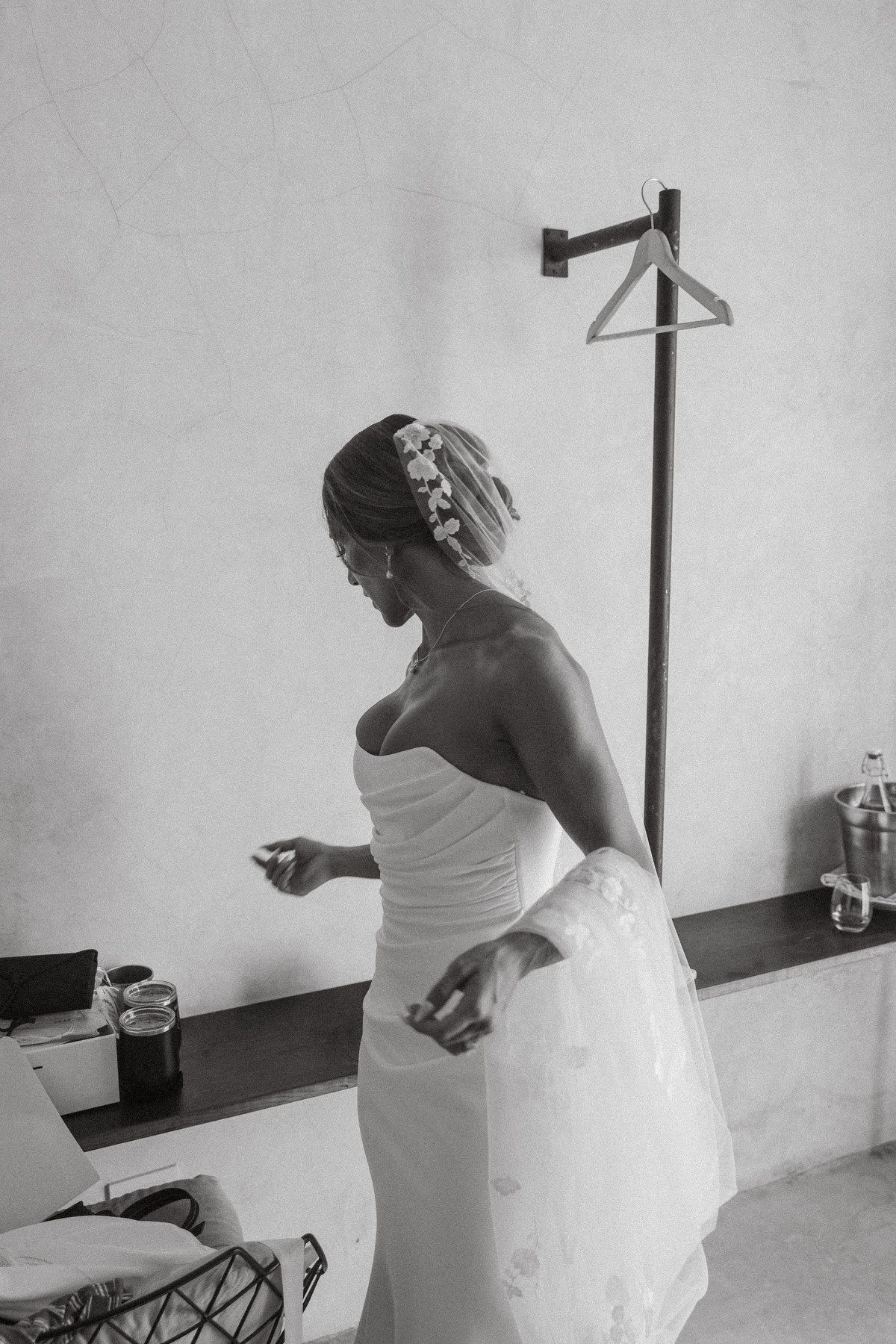 Black and white photo of a bride in a strapless wedding gown with floral hair accessories getting ready indoors.