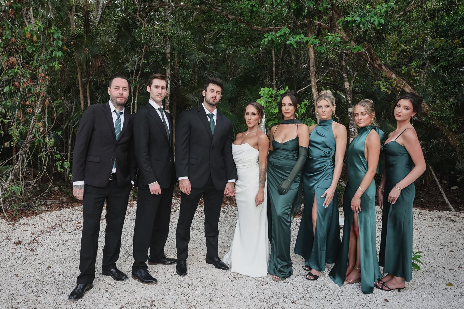 Wedding party poses outdoors in lush greenery, groomsmen in black suits and bridesmaids in emerald green satin dresses.