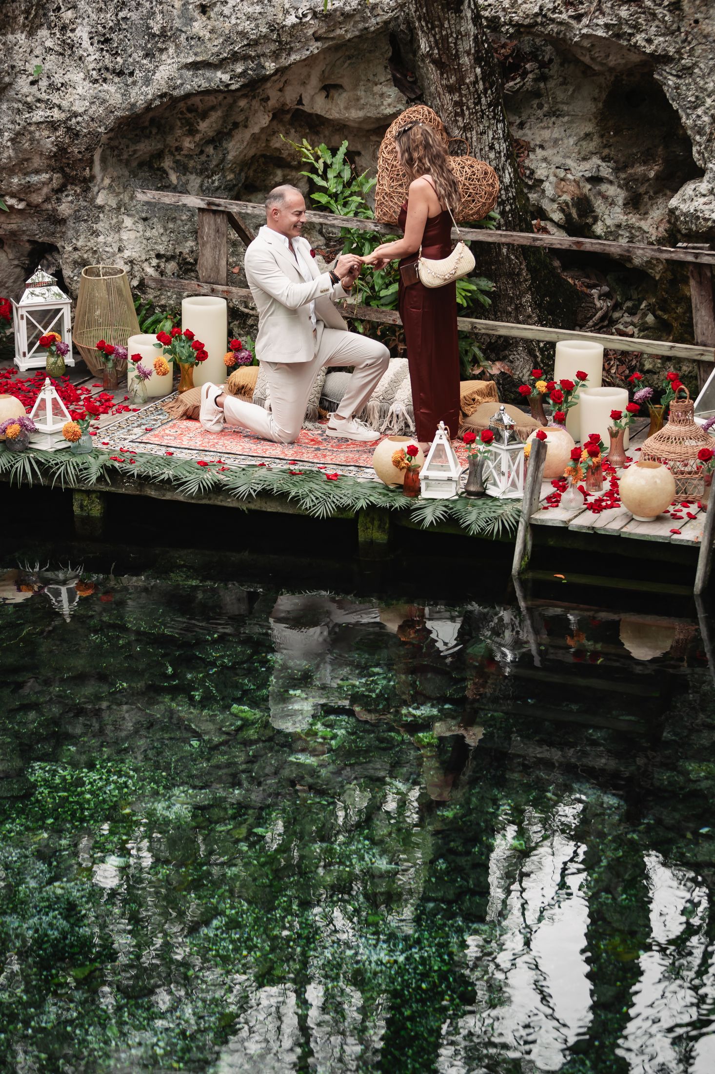 Man proposes to woman on romantic candlelit dock over crystal clear cenote, surrounded by red roses and lanterns.