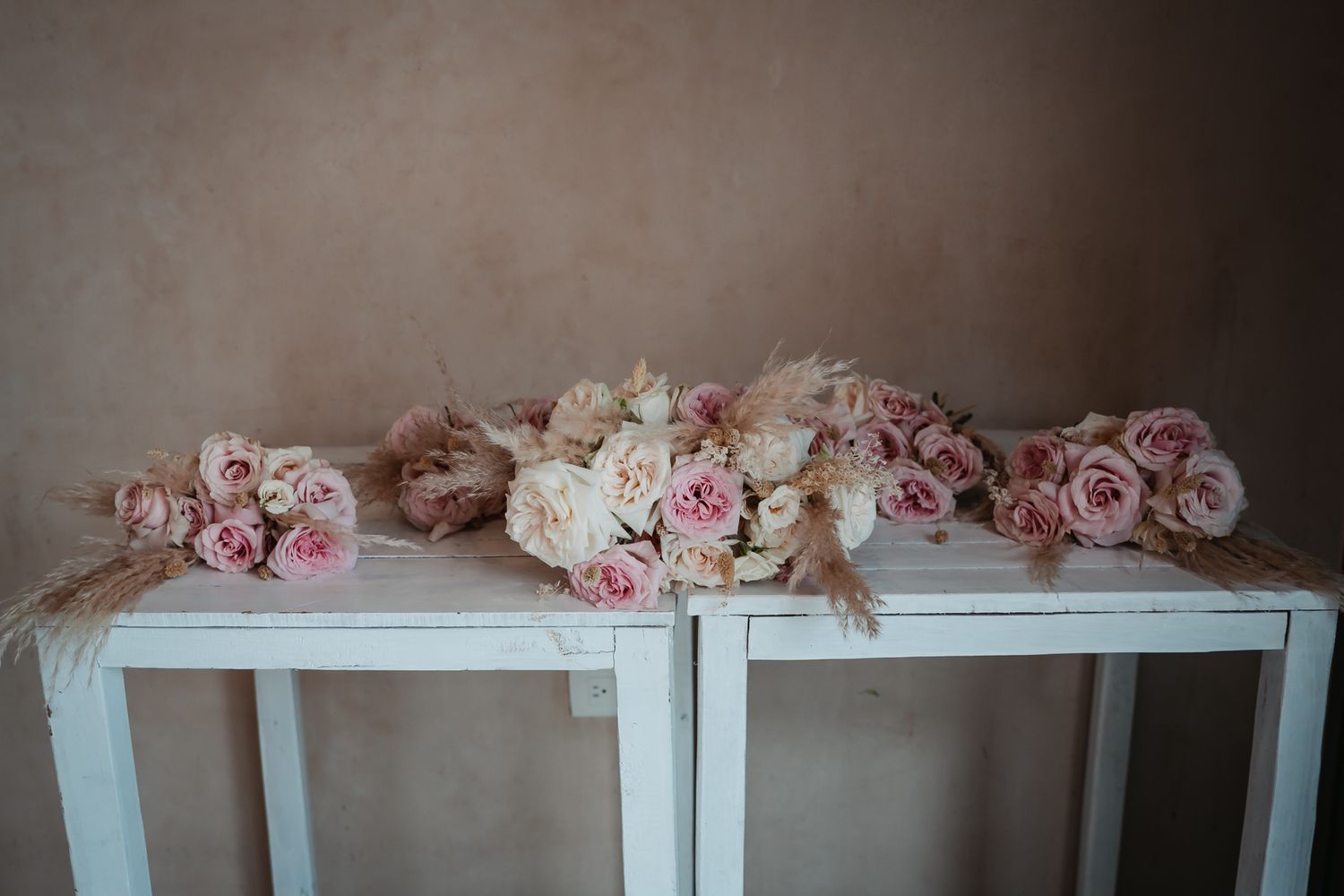 Elegant arrangement of pink and cream roses with pampas grass displayed on a white table against a beige wall.