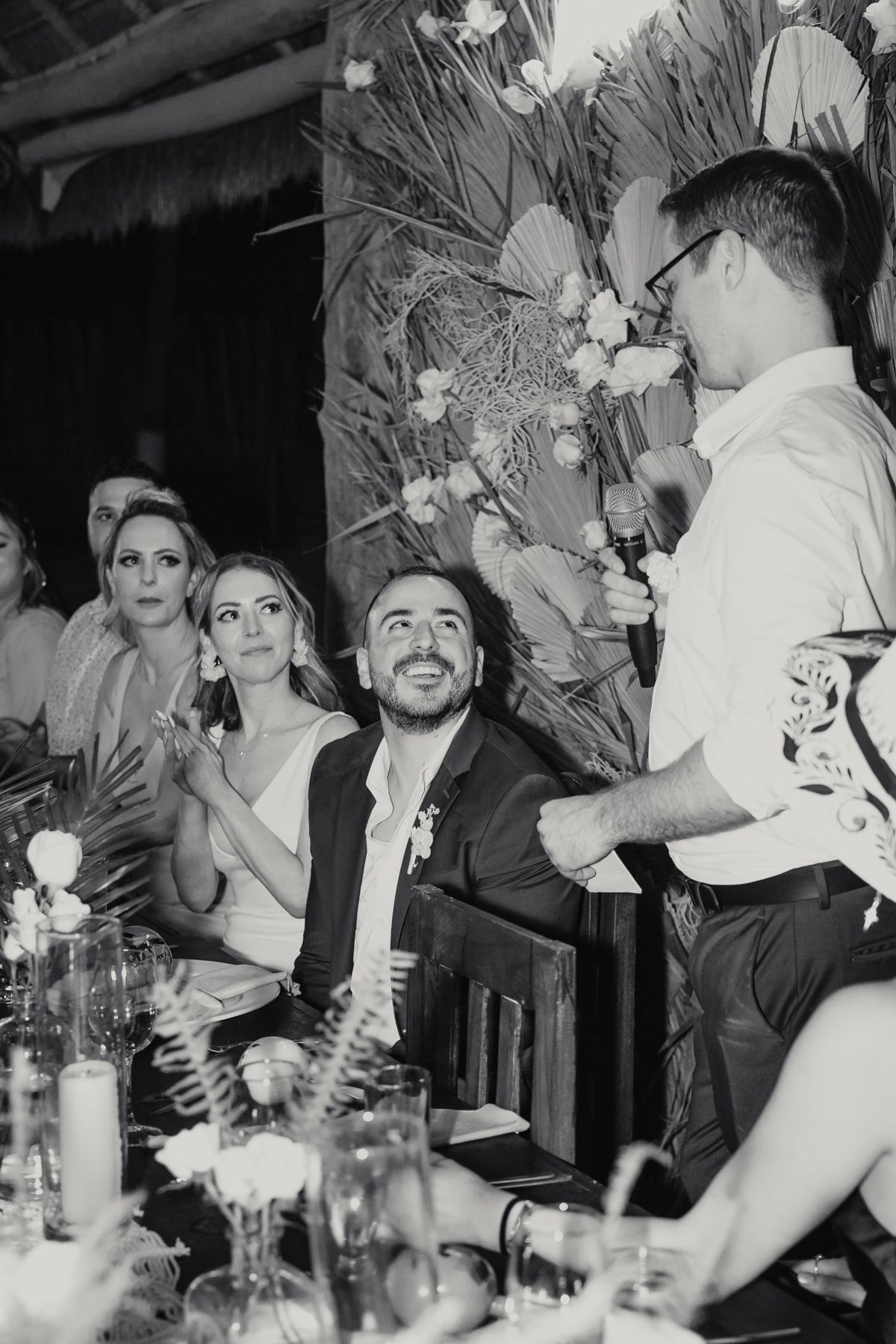 Black and white wedding reception photo of bride and groom laughing during a heartfelt speech.
