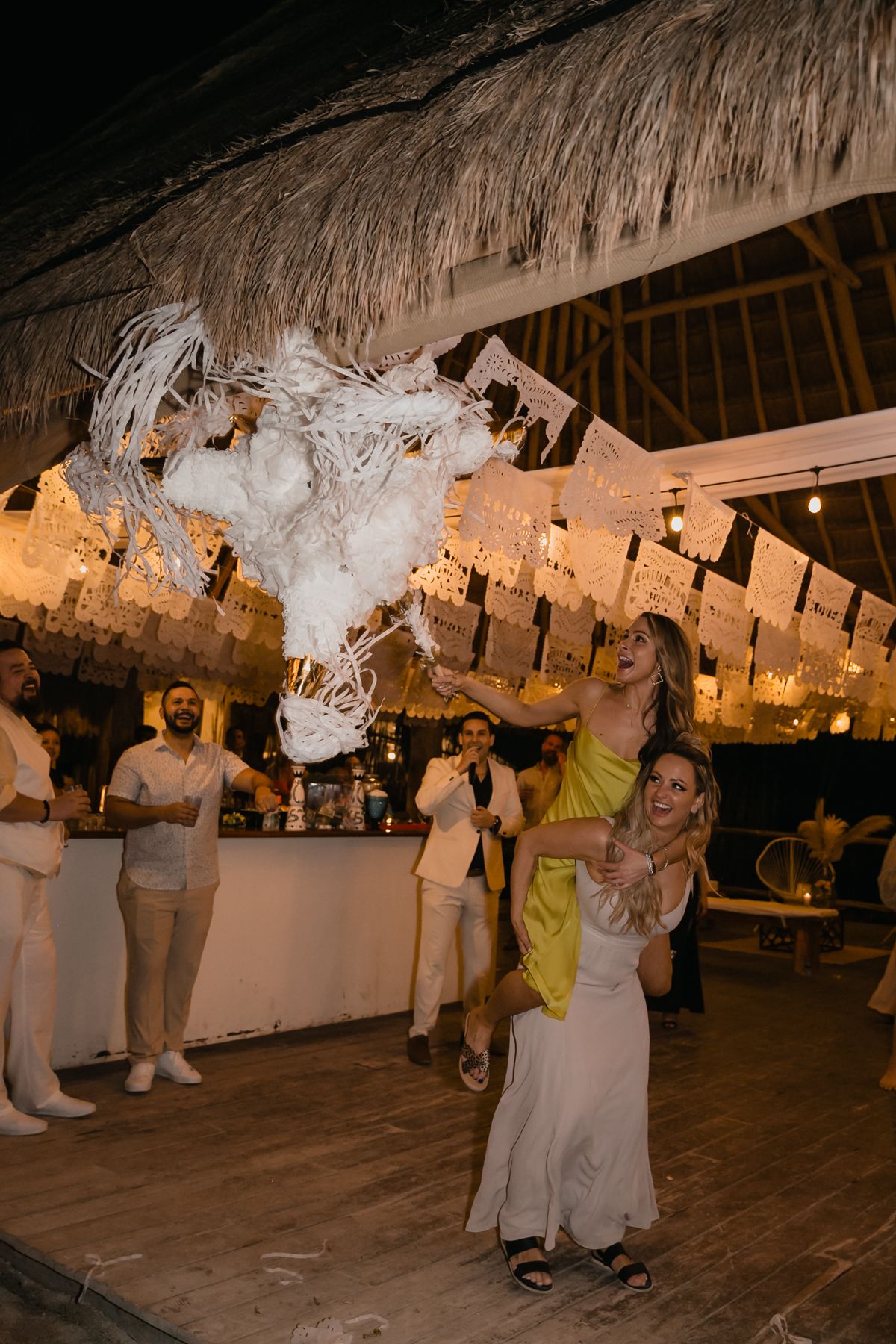 Bride hits white pinata under thatched roof decorated with papel picado banners at Mexican wedding reception.