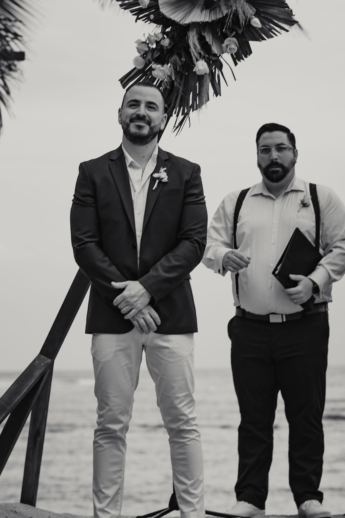 Groom smiling at altar during beach wedding ceremony with officiant holding book beside him.