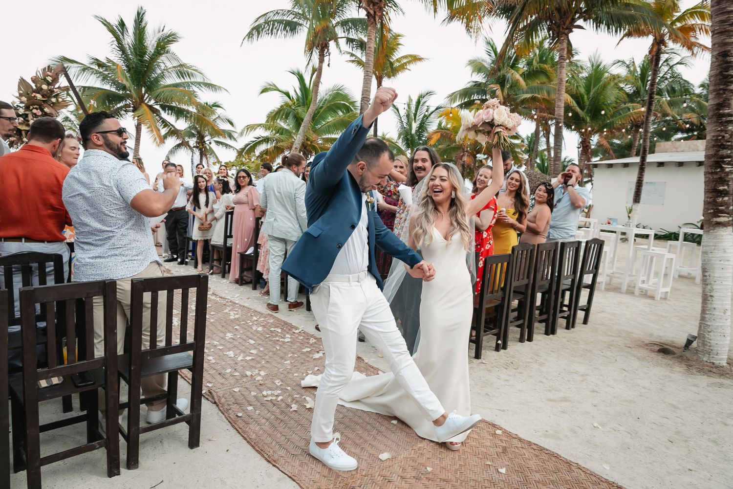 Joyful newlyweds celebrate recessional walk among cheering guests at tropical beach wedding.
