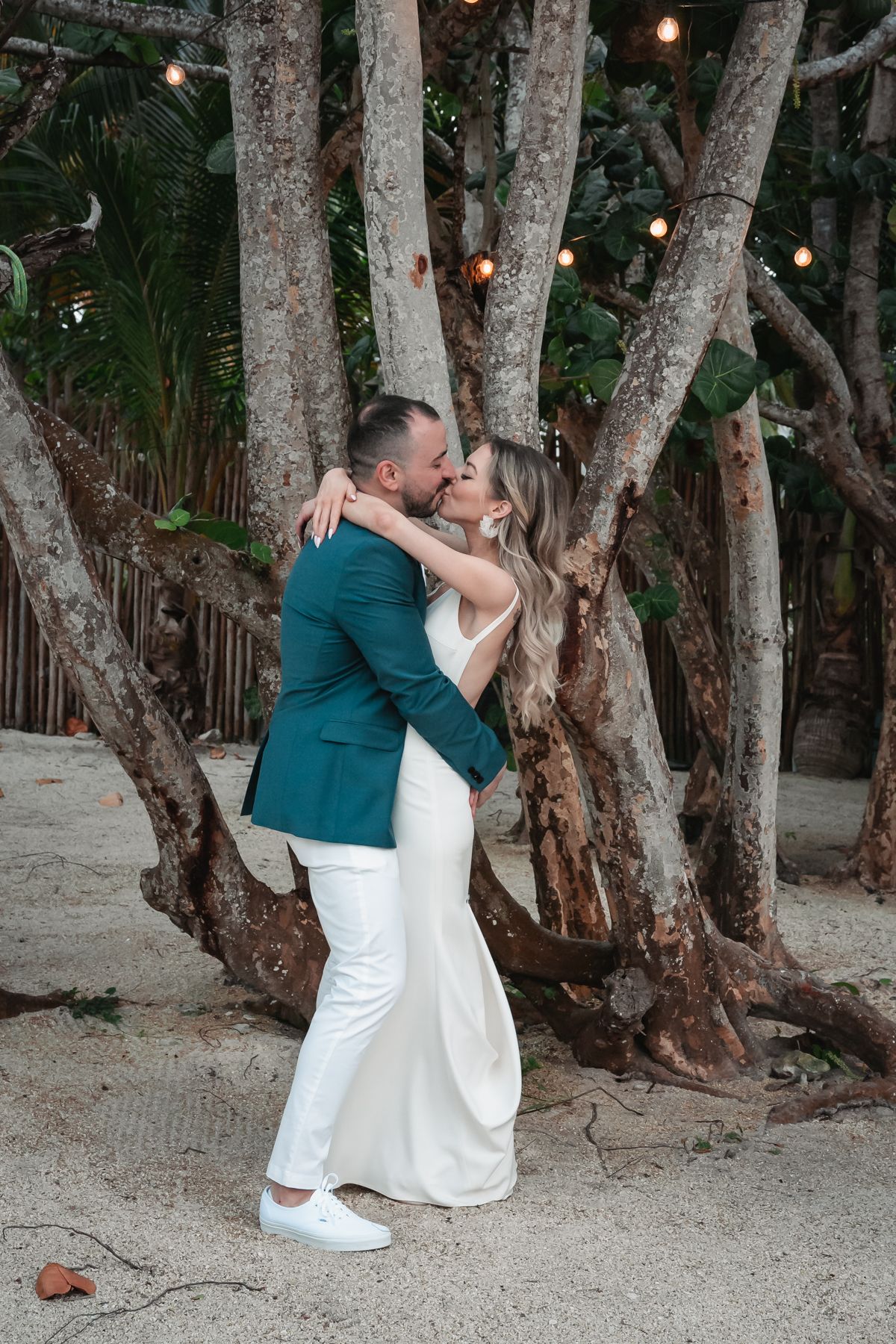Bride and groom share a romantic kiss under string lights among tropical trees on beach.