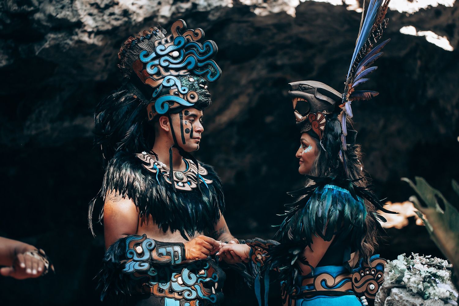 Two people in elaborate Mayan ceremonial costumes with turquoise and black feathered headdresses in a cave setting.