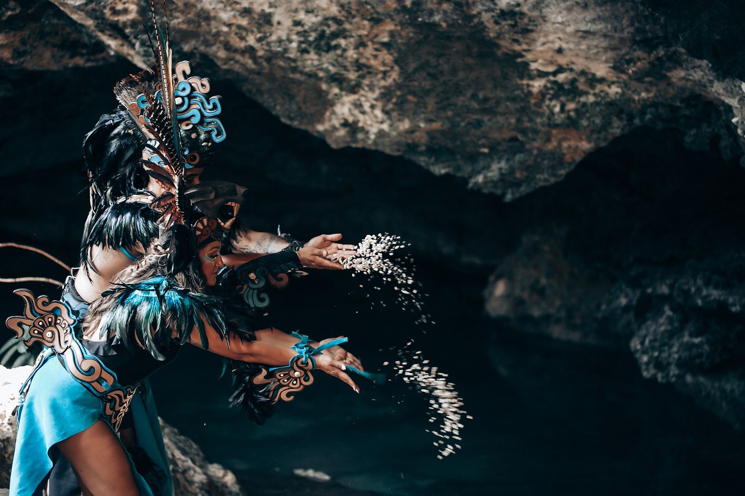 Performer in Mayan ceremonial blue costume extends hand dramatically near a dark cave opening.