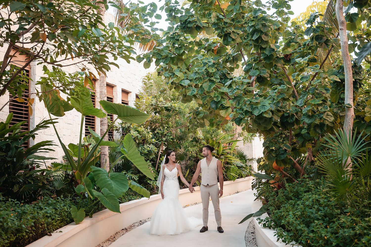 Bride and groom hold hands walking along a lush tropical garden path at an outdoor wedding venue.