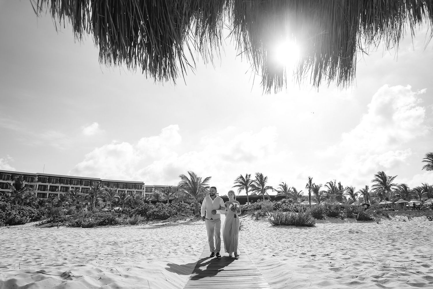 Couple standing on white sandy beach in black and white photo with palm trees and resort in background.