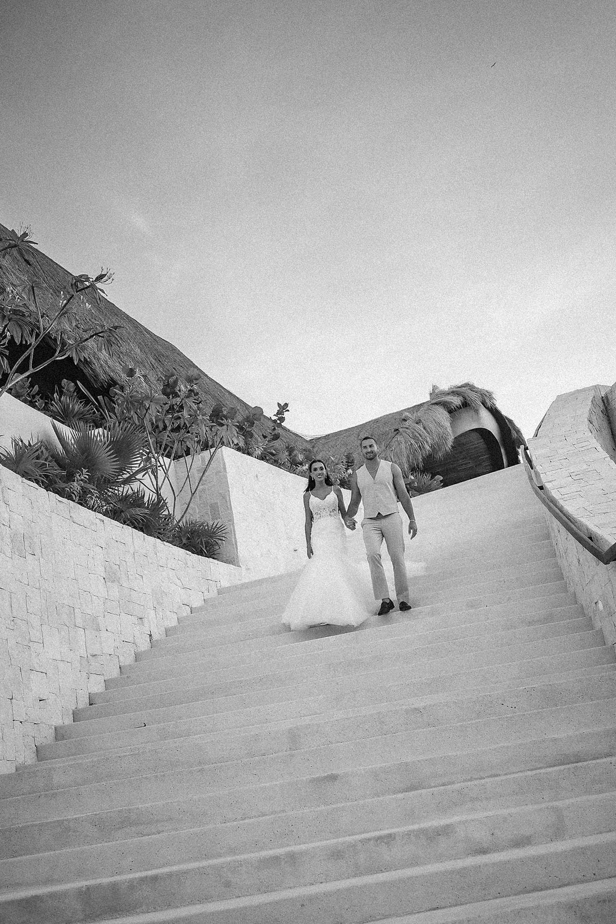 Black and white photo of a bride and groom walking down grand outdoor stairs at a destination wedding venue.