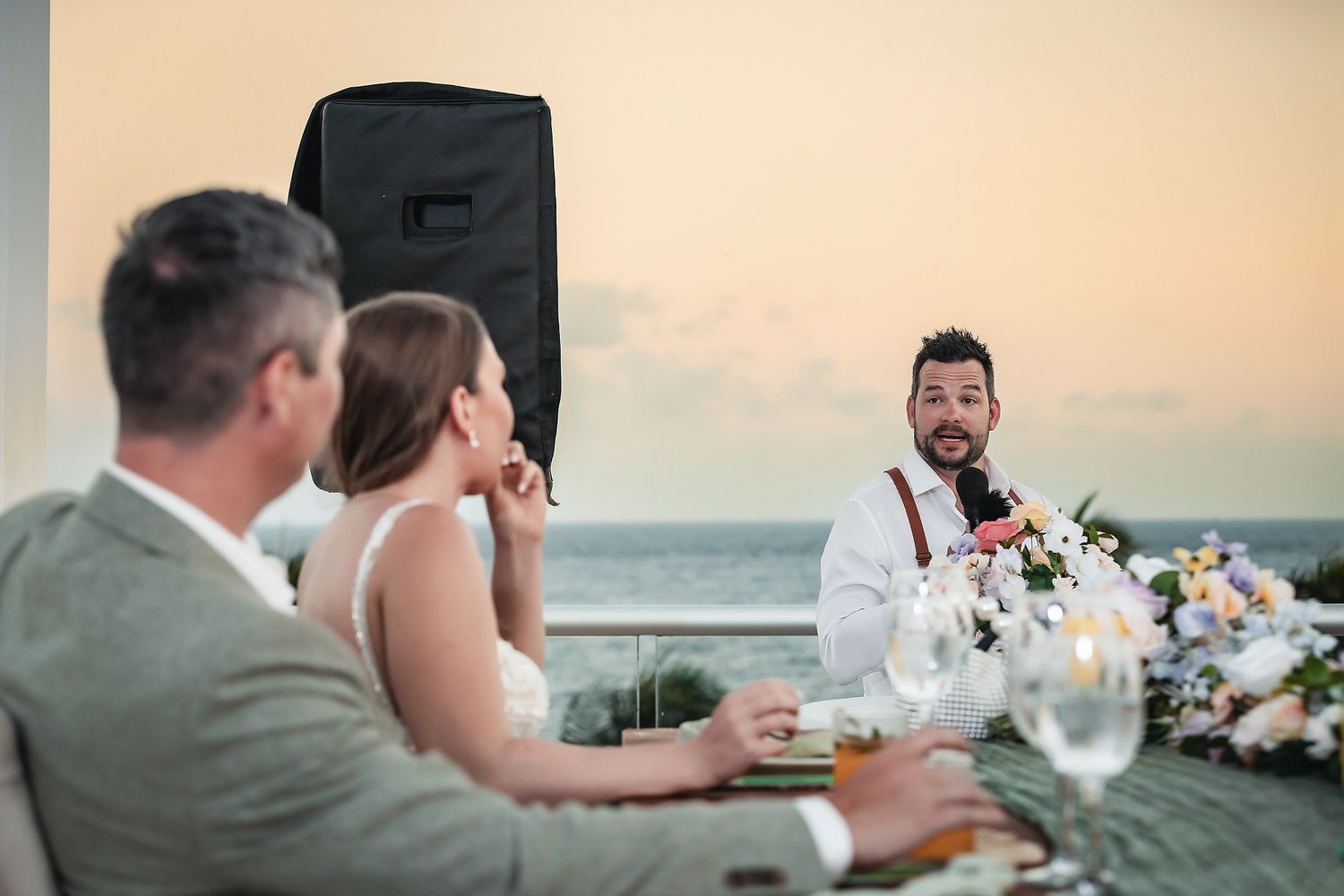 Groom giving a speech at an outdoor beachside wedding reception at sunset, with bride and guests seated at table.
