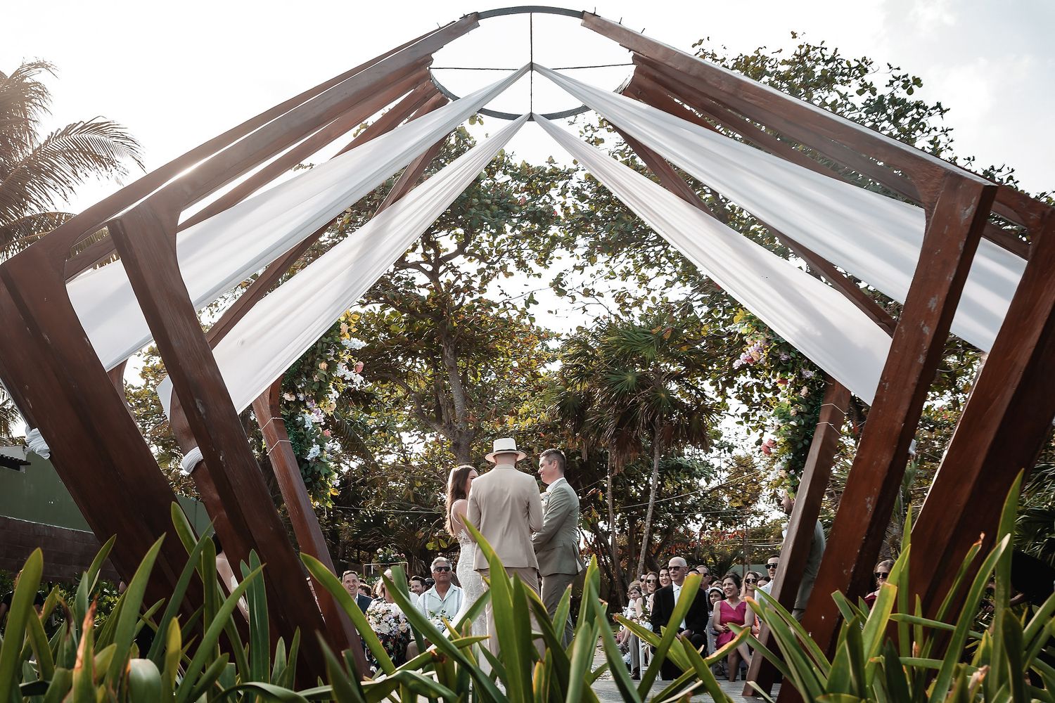 Outdoor wedding ceremony under wooden arch with white draping, surrounded by lush greenery and guests.
