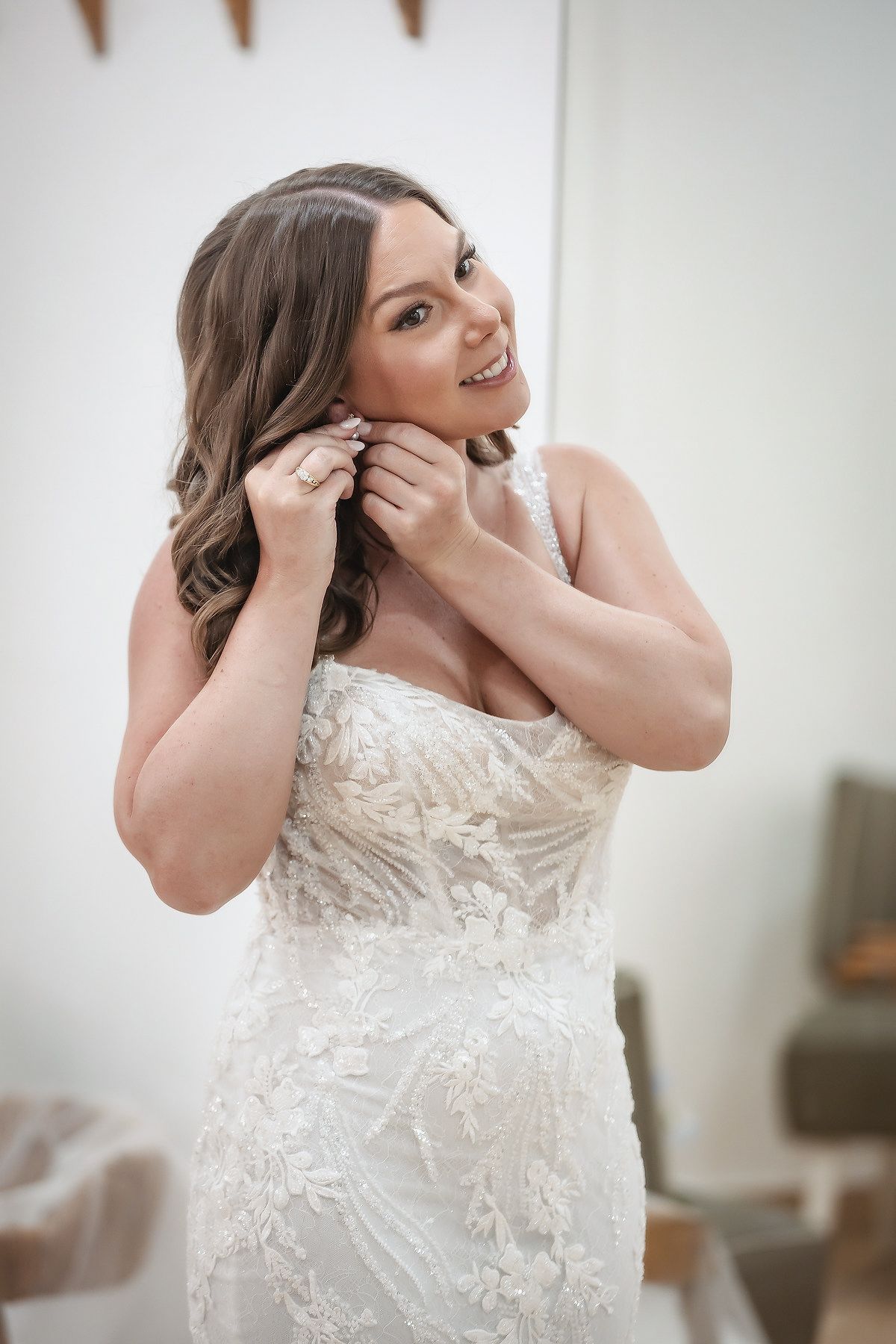 Smiling bride in a strapless lace wedding gown putting on earrings while getting ready for her wedding day.