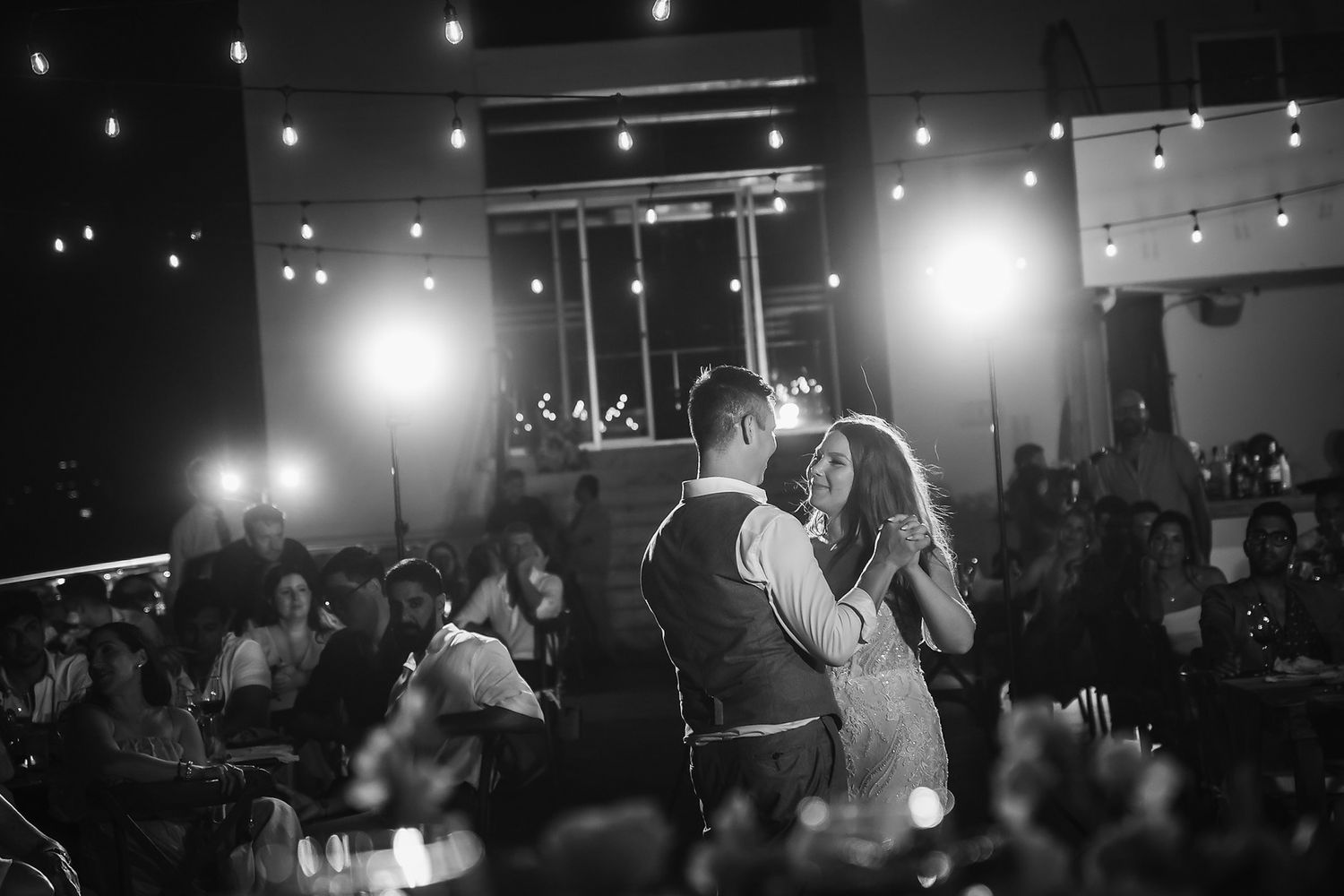Black and white photo of a couple sharing their first dance at a wedding reception with guests watching.