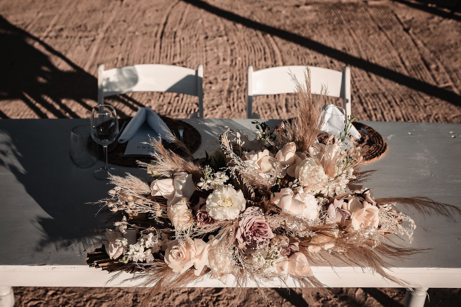 Elegant boho wedding bouquet with dried pampas grass and blush roses resting on white chairs at outdoor ceremony.