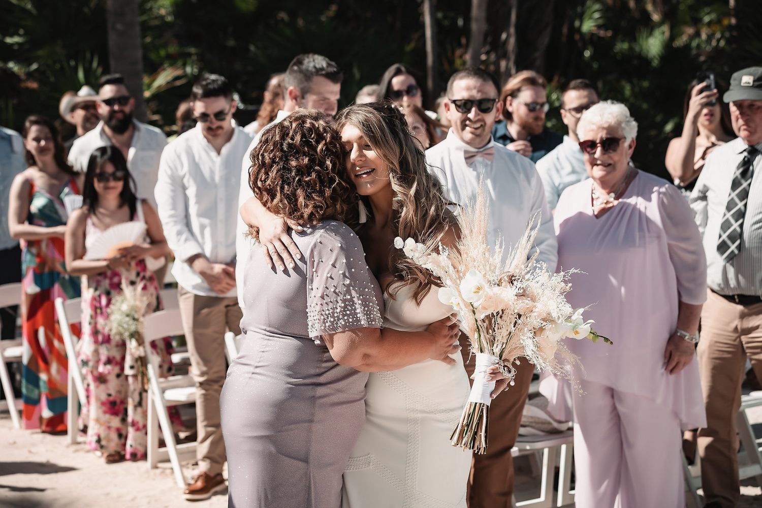 Bride hugging a guest at an outdoor wedding ceremony while holding a dried floral bouquet.