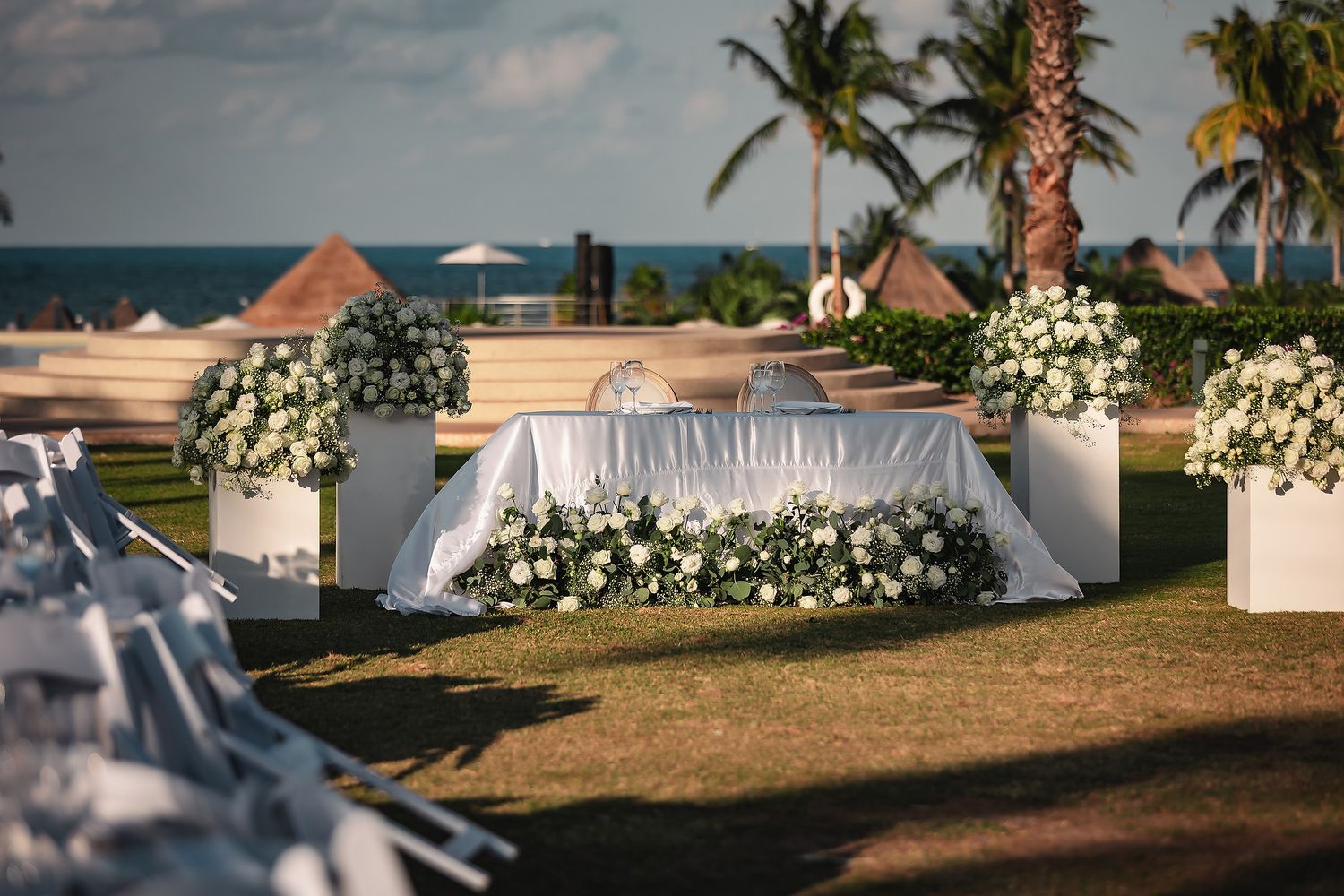 Elegant outdoor wedding setup with white floral arrangements, draped table, palm trees, and ocean view at a tropical resort.
