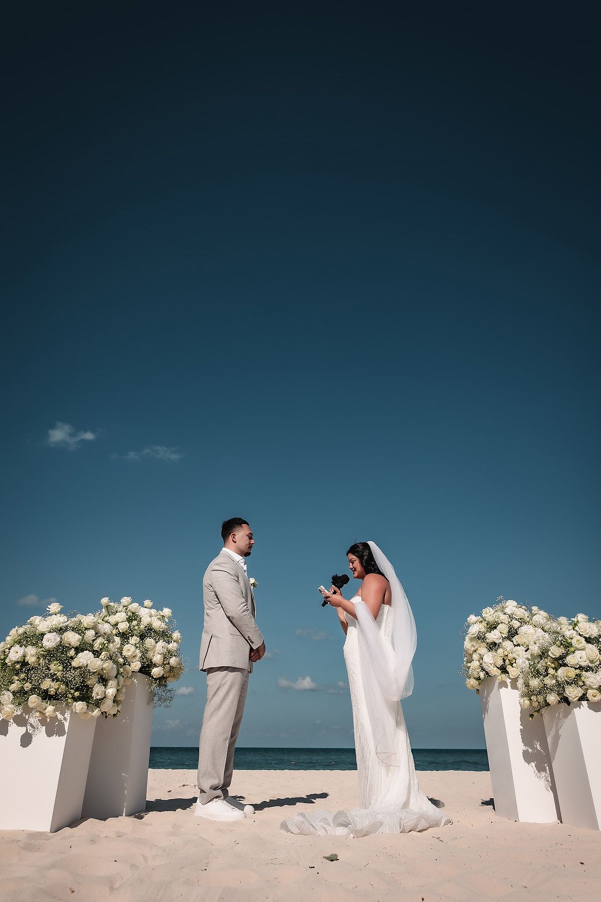 Bride and groom exchange vows on a sandy beach altar adorned with white floral arrangements under a clear blue sky.