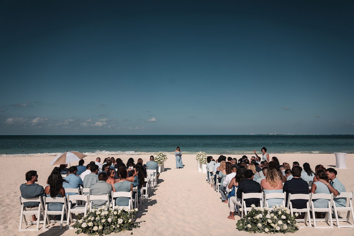 Outdoor beach wedding ceremony with guests seated in white chairs facing the ocean altar.