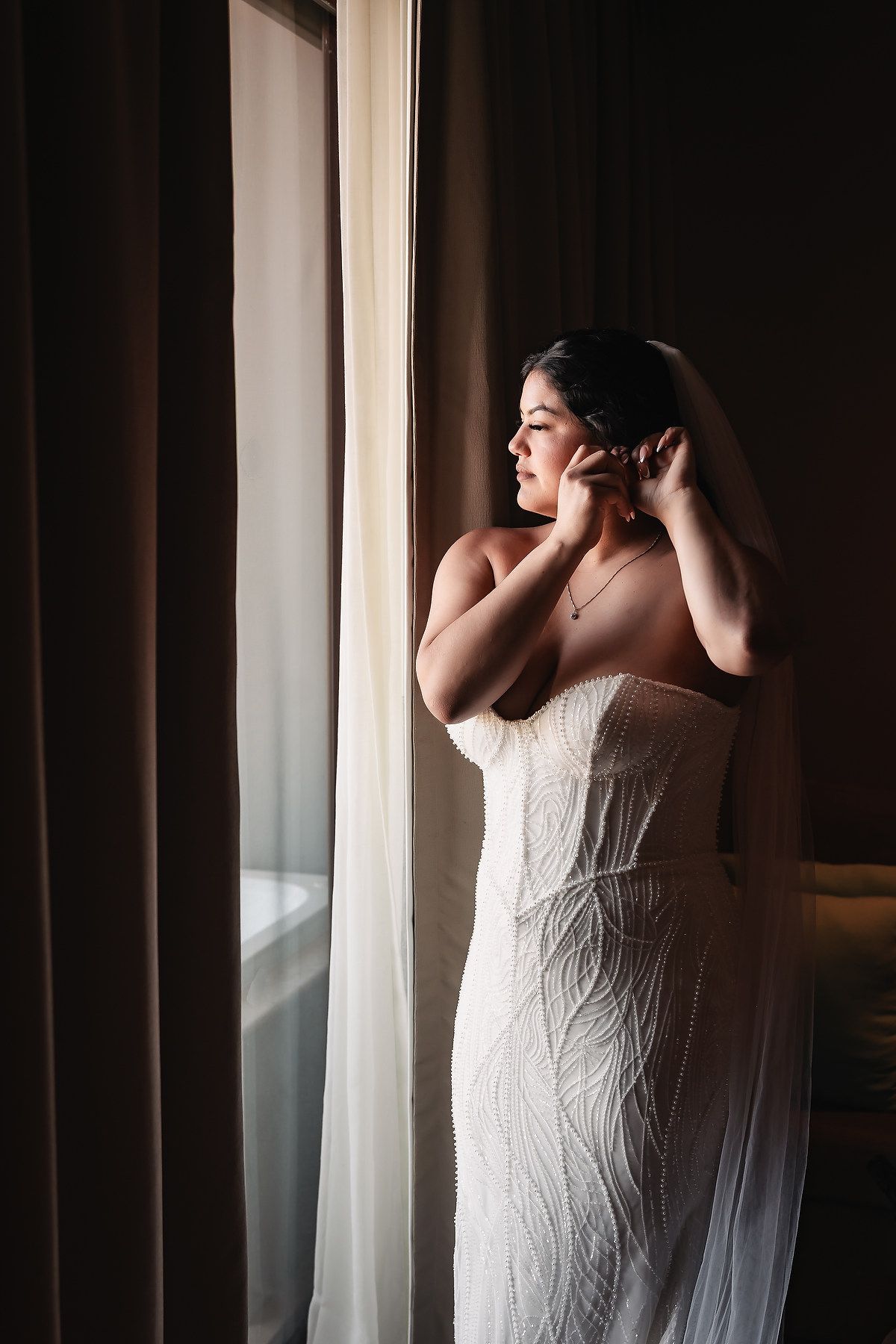 Bride in a white lace strapless gown adjusting her earrings by a window with soft natural light.