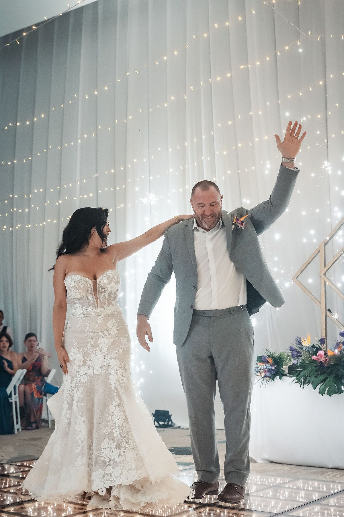 Bride in lace gown and groom in gray suit celebrate their first dance at a wedding reception with twinkling lights.