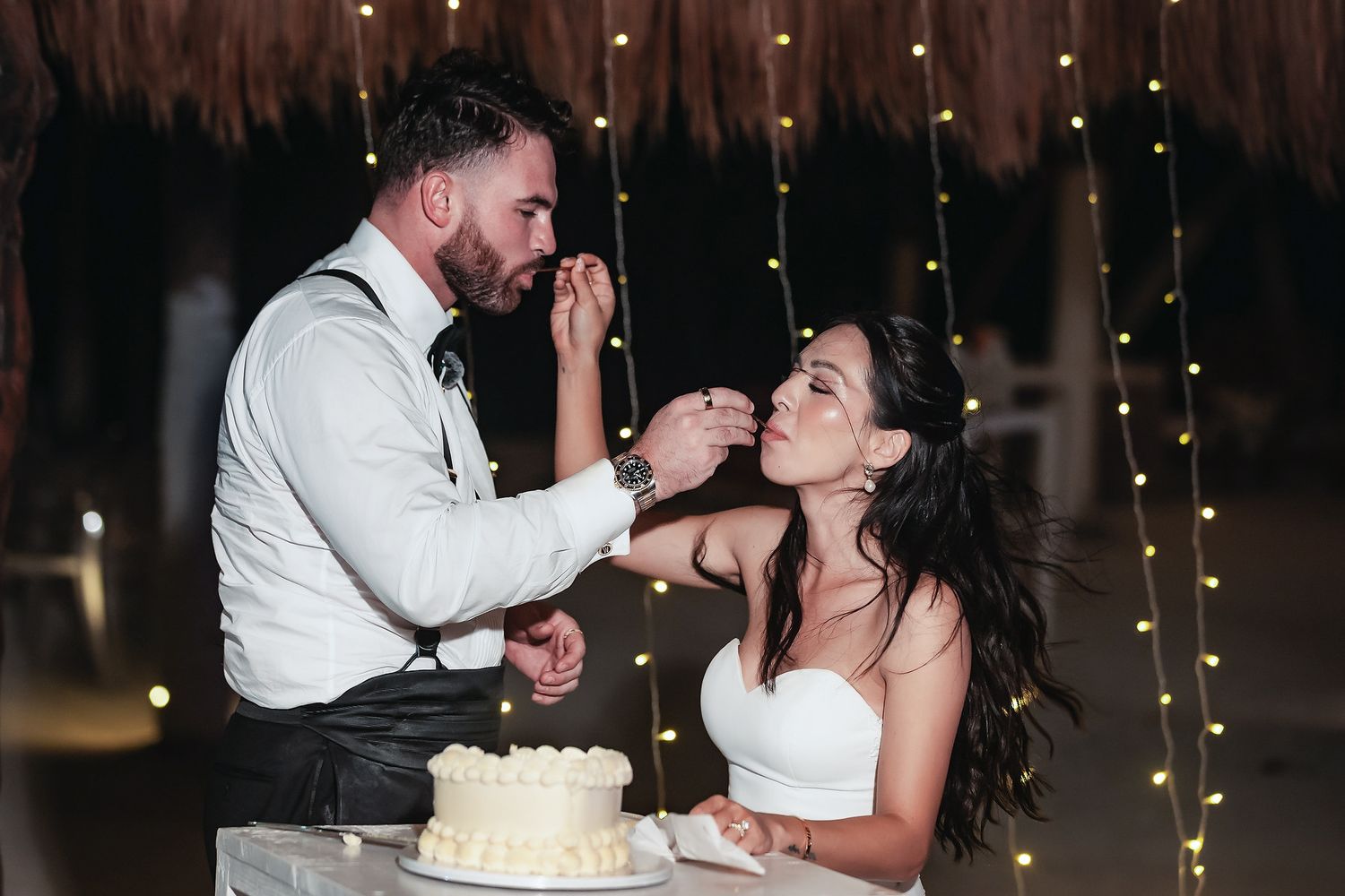 Bride and groom feeding each other wedding cake at a romantic reception with fairy lights in the background.