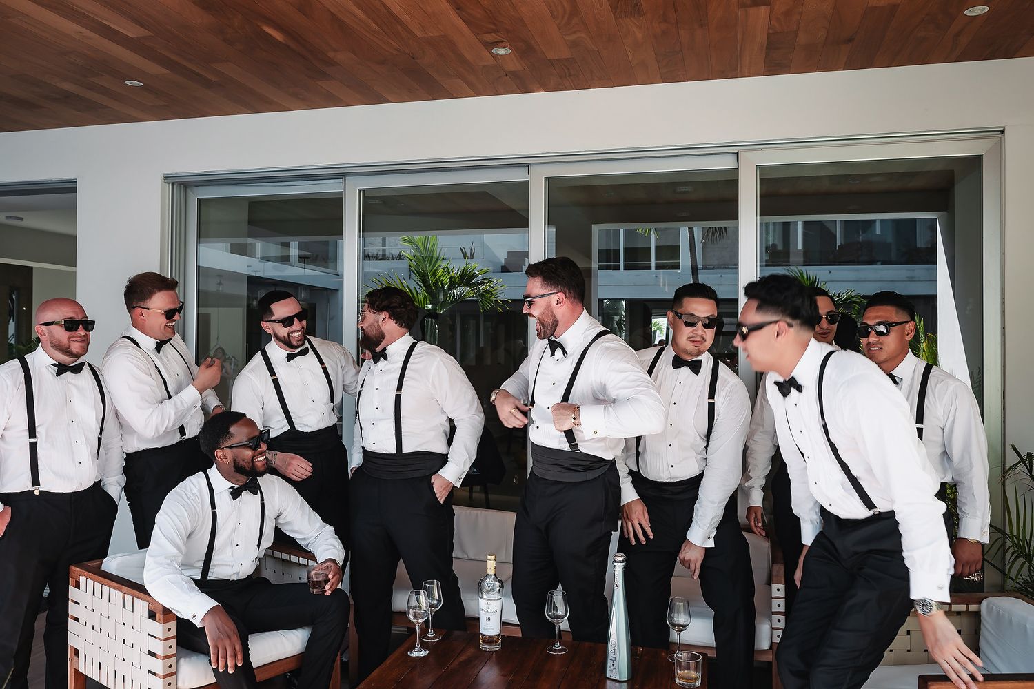 Groomsmen in white shirts, black suspenders, and bow ties celebrate together on a modern outdoor patio before a wedding.