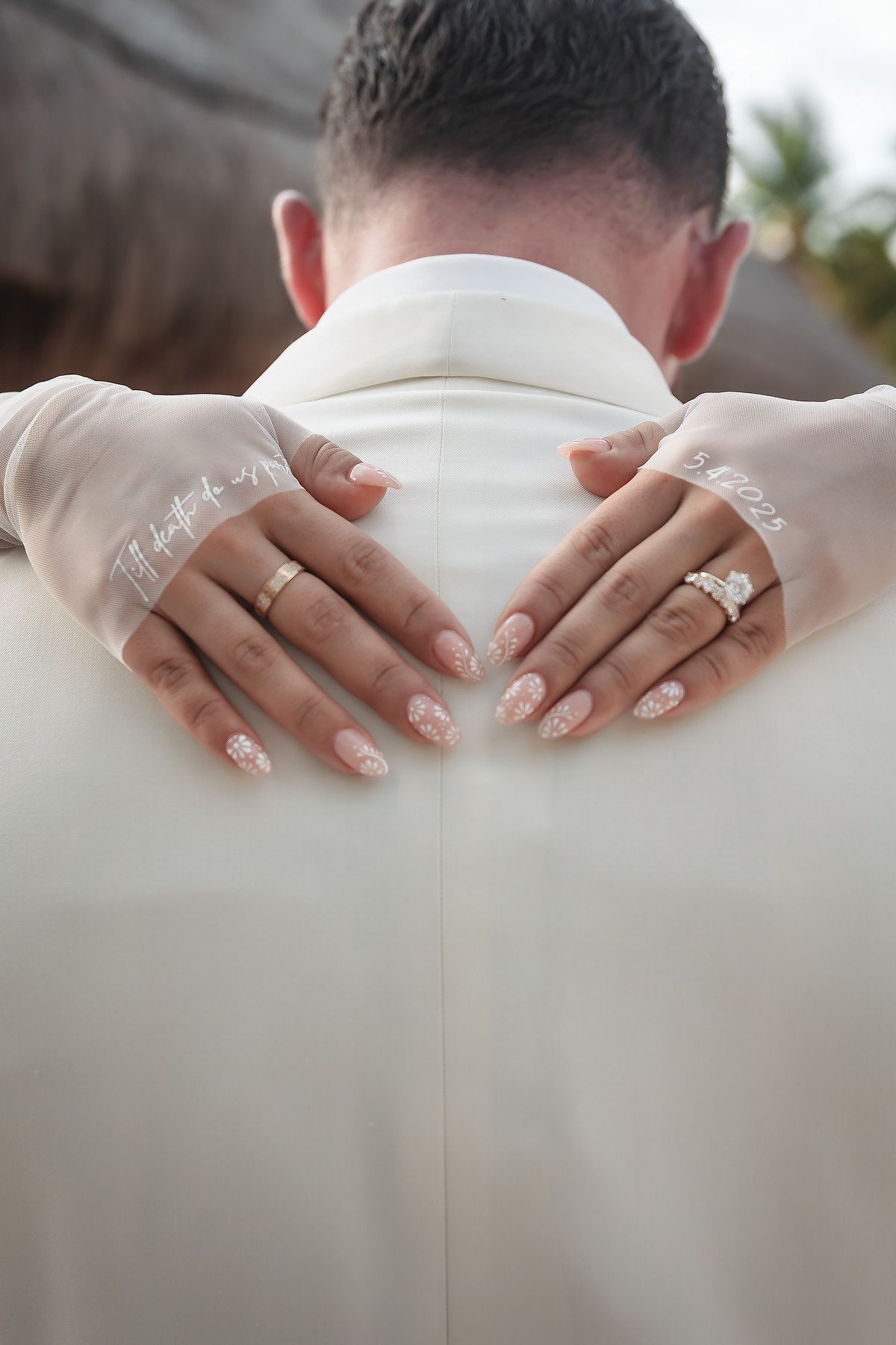 Bride's hands with elegant lace gloves and diamond ring resting on groom's white suit jacket at wedding.