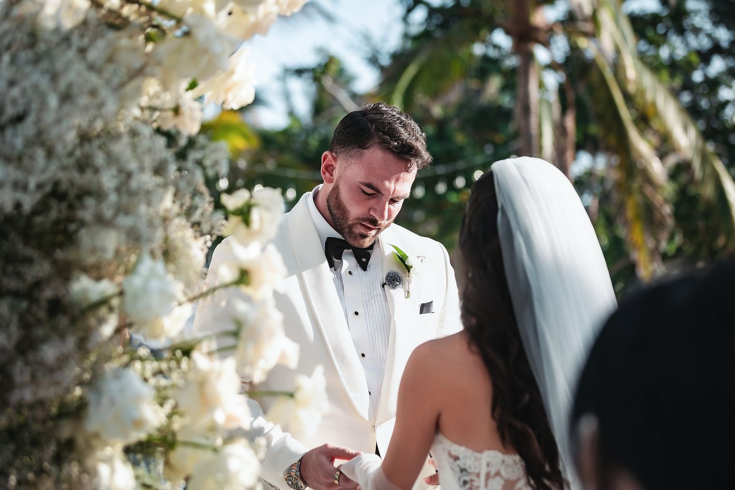 Groom in white tuxedo exchanges vows with bride at outdoor wedding ceremony surrounded by white floral arrangements.