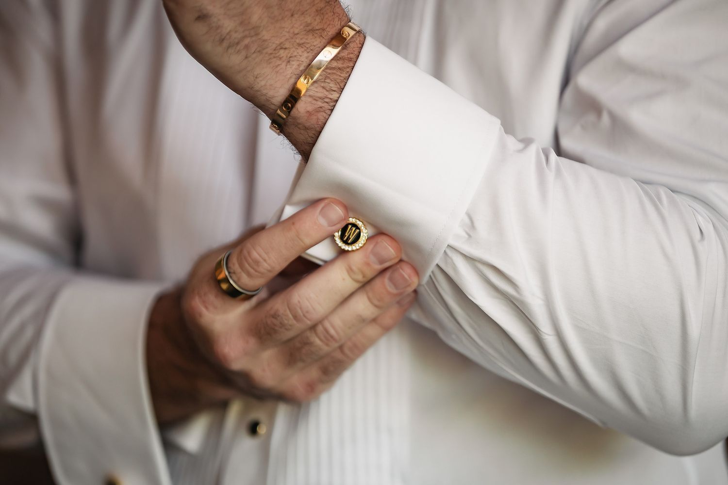 Man in white dress shirt fastening gold cufflinks, wearing gold bracelet and signet ring, formal wedding attire.