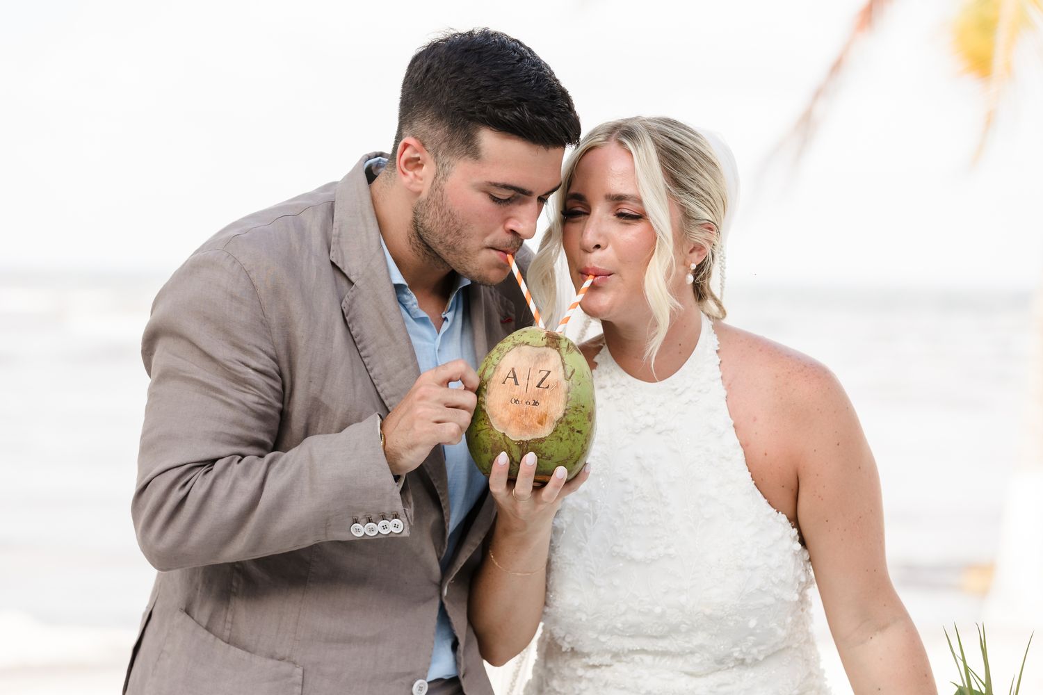 Couple in wedding attire sharing a coconut drink on a beach, symbolizing a romantic tropical celebration.