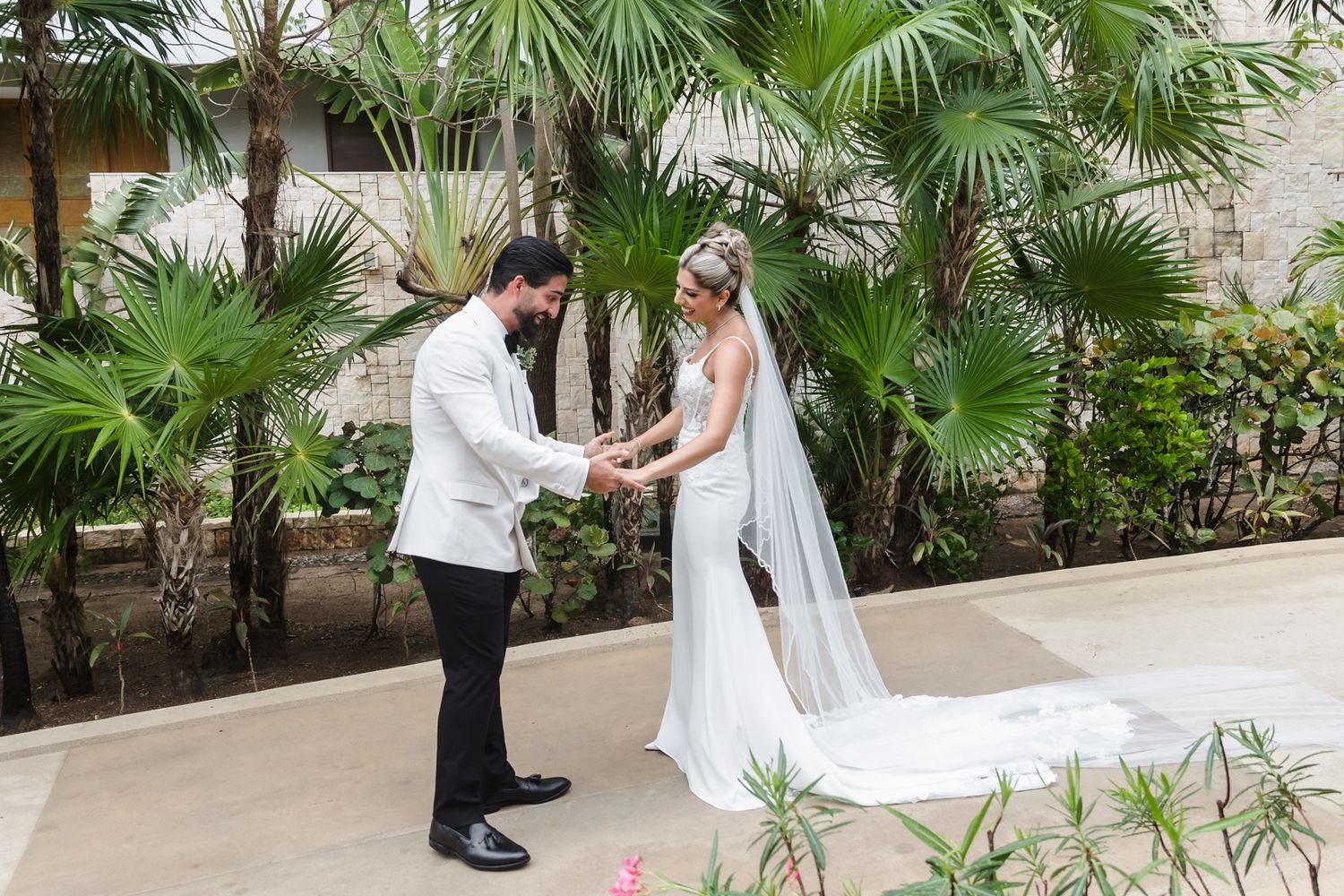 Bride and groom hold hands facing each other in a tropical garden, bride wearing a flowing white gown with veil.