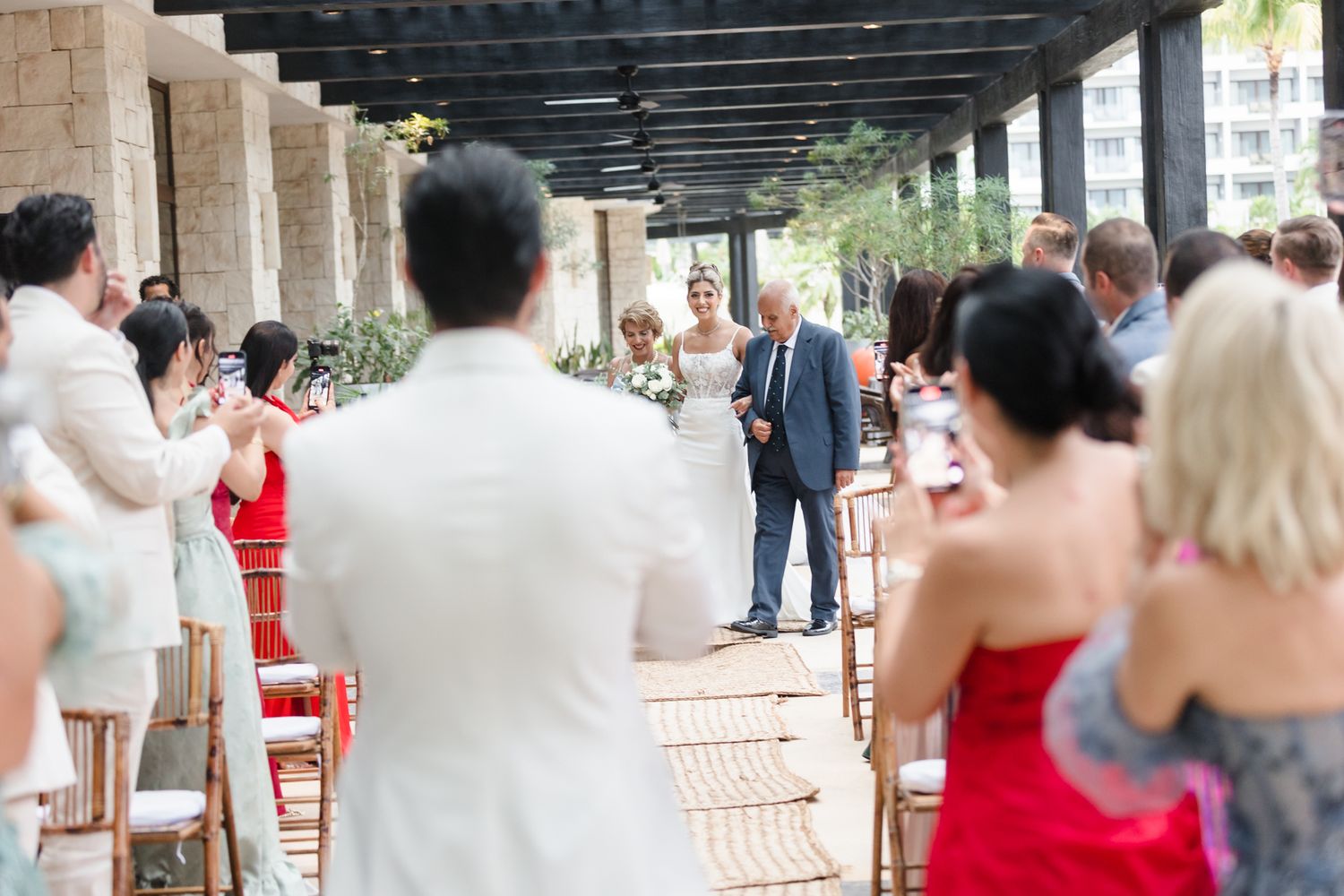Bride walks down the aisle with her father at an outdoor wedding ceremony while guests applaud and capture photos.