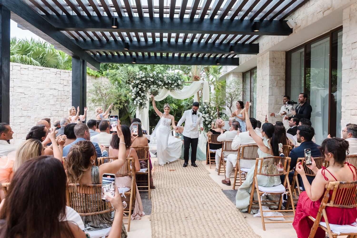 Bride and groom celebrate at outdoor wedding ceremony under pergola with seated guests in tropical venue.