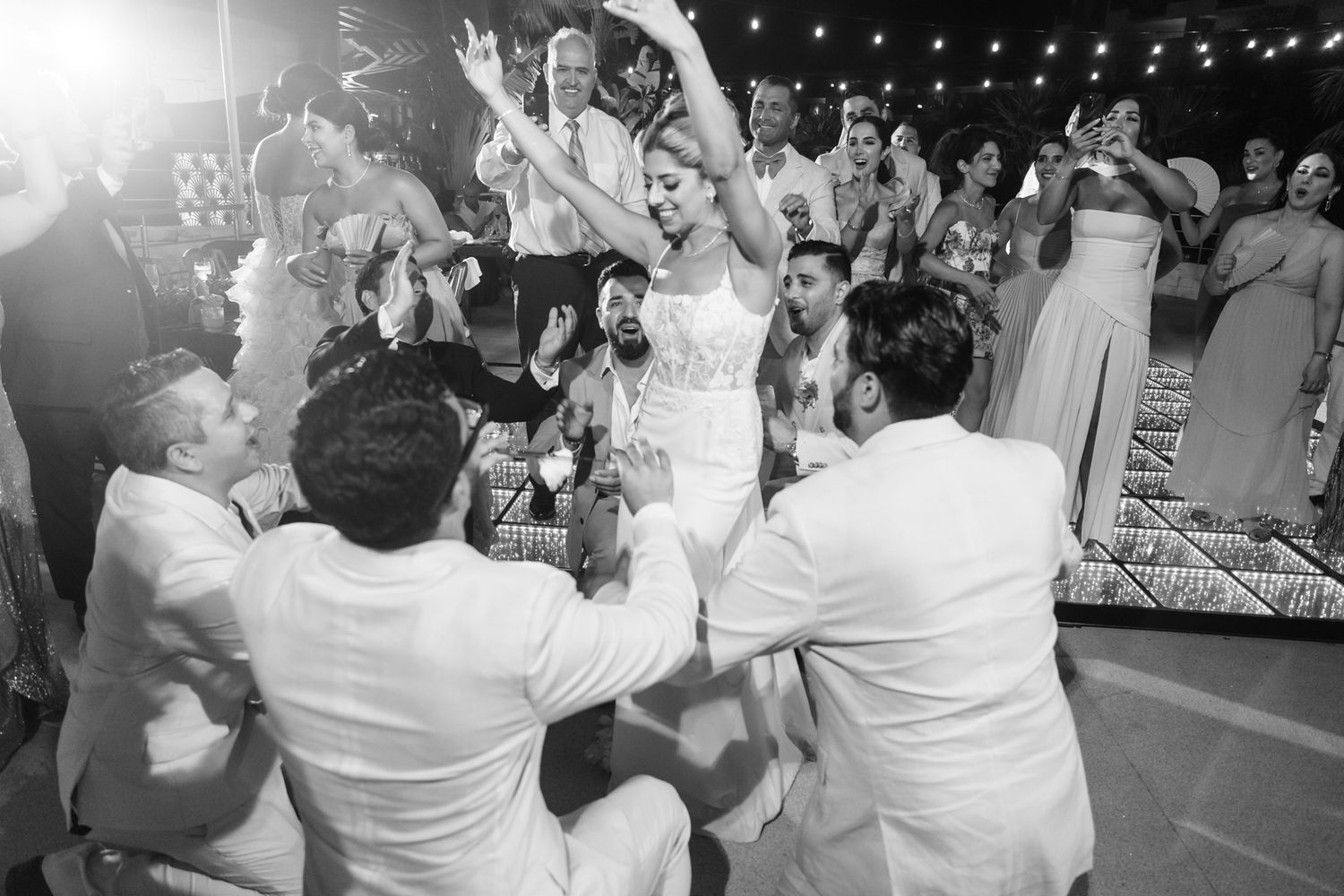 Black and white wedding reception photo of bride dancing joyfully surrounded by guests in formal attire.