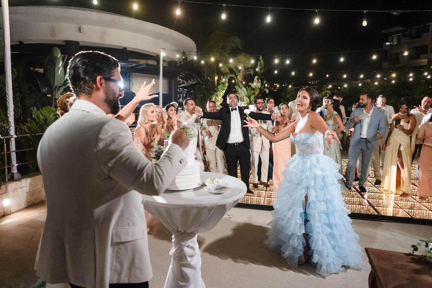 Bride in blue ruffled gown and groom in white suit celebrate at outdoor wedding reception with guests and string lights.