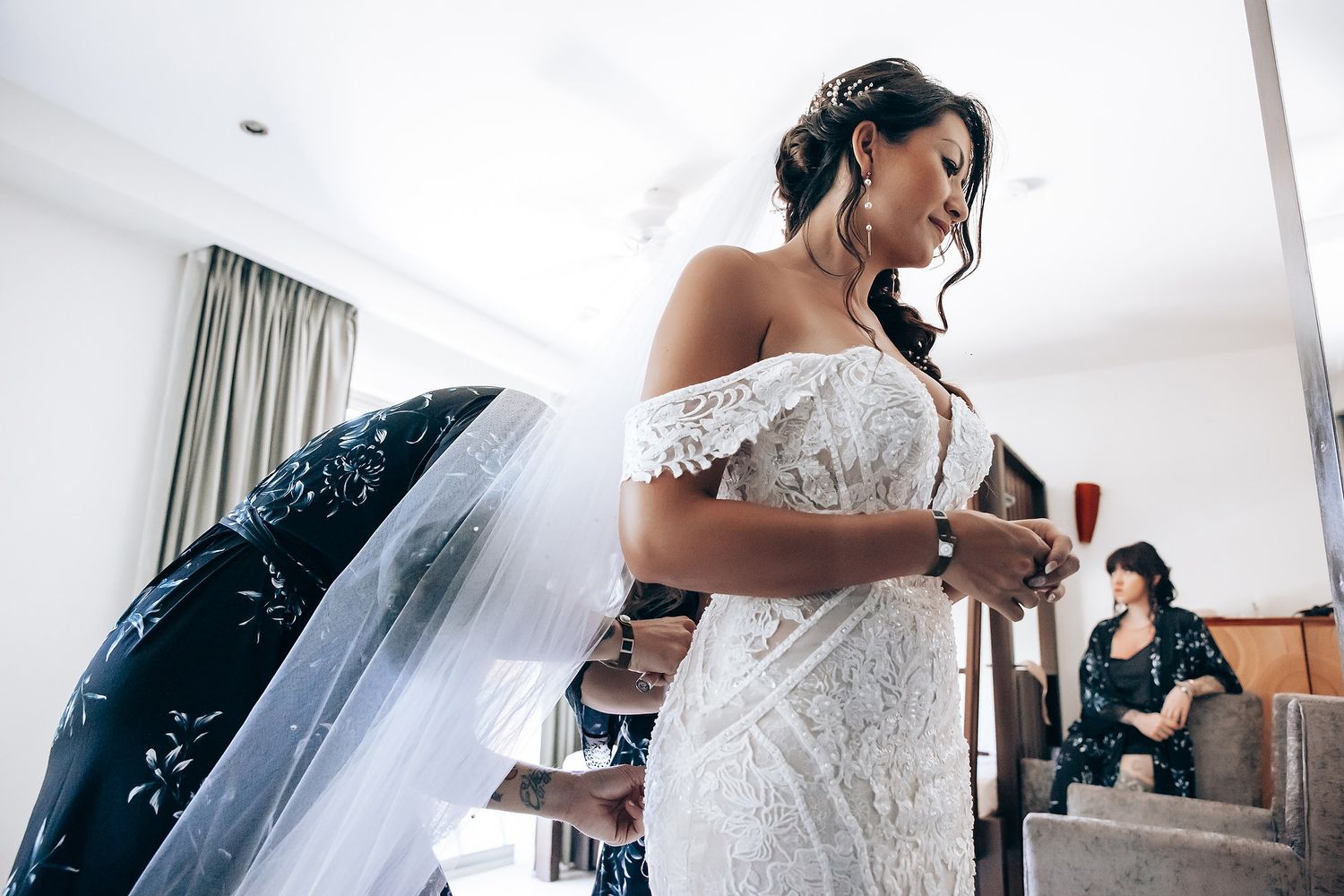 Bride in an elegant off-shoulder lace wedding dress getting ready in a bright hotel room.