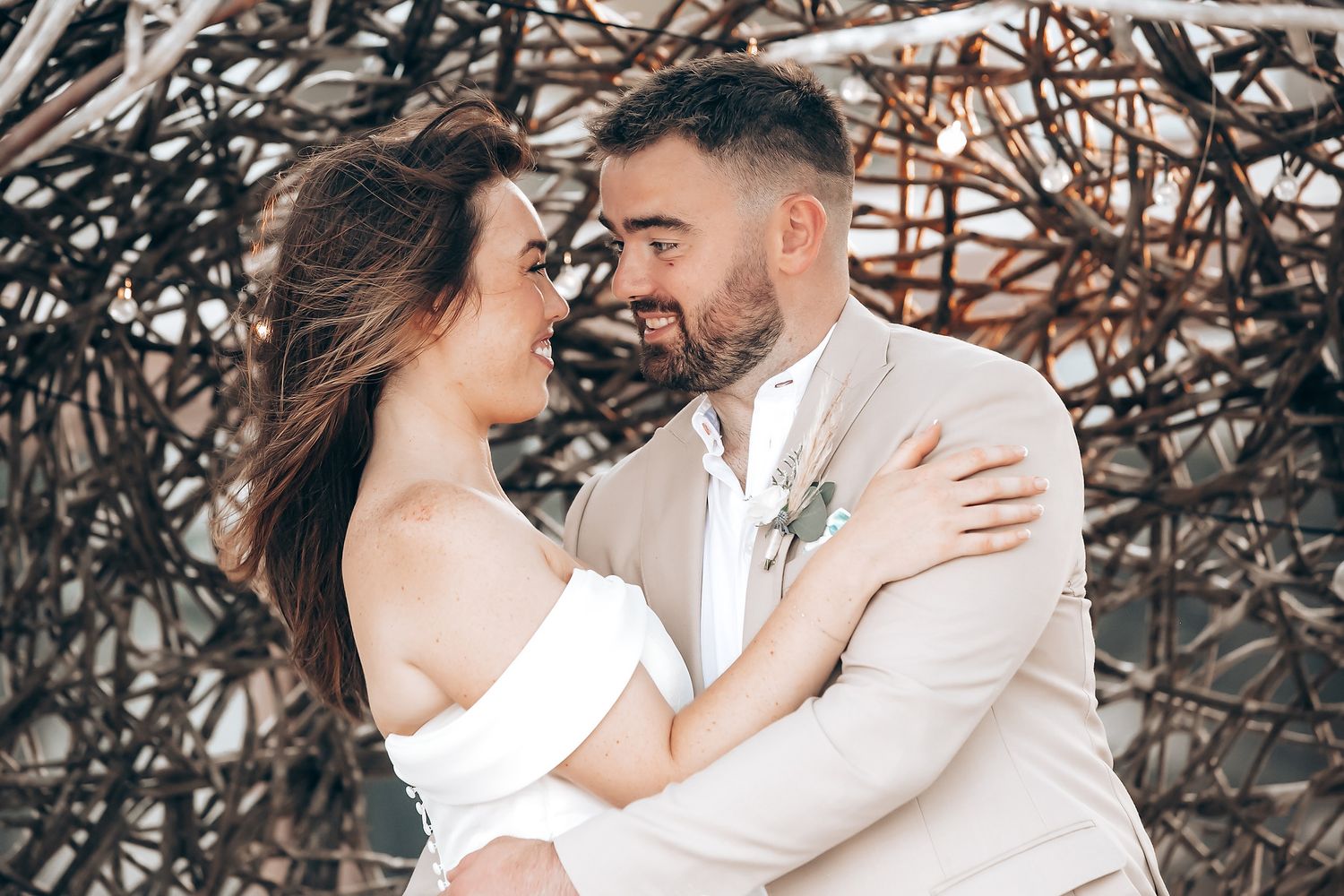 A romantic wedding couple embraces outdoors against a natural branch backdrop in soft neutral tones.