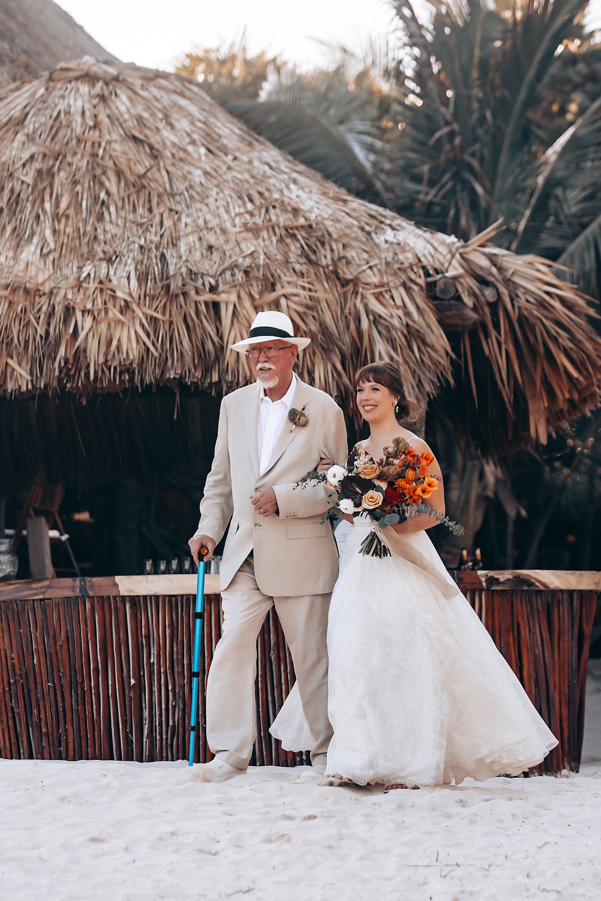 Beach wedding couple poses under thatched palm roof on white sand.