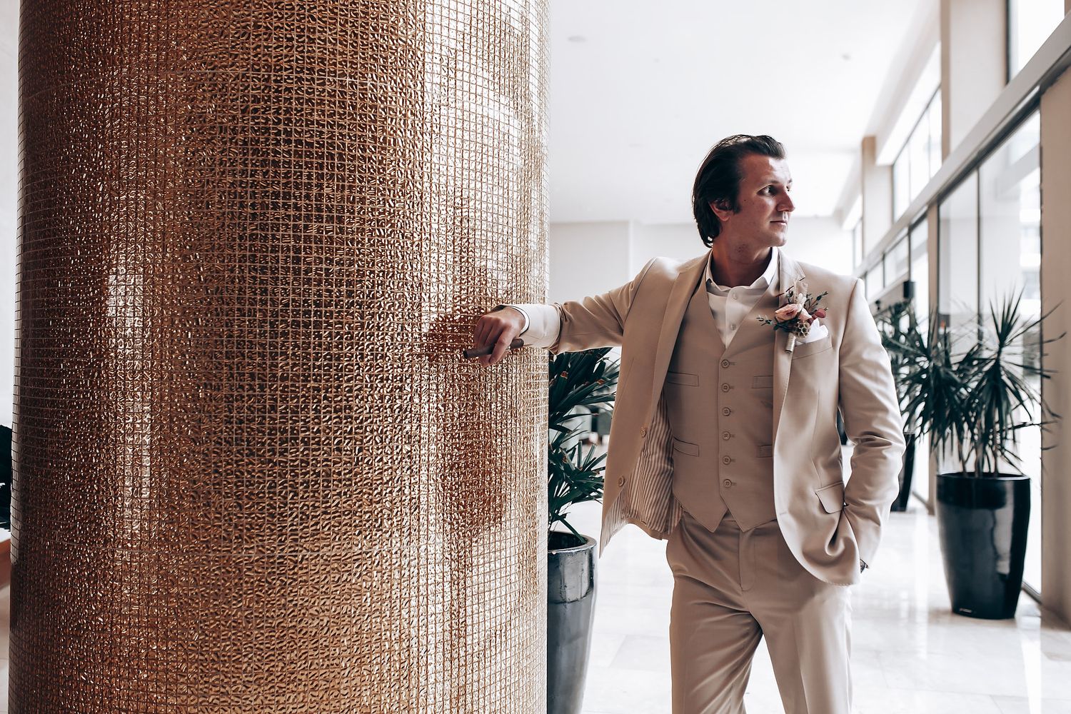 A person in a light beige suit stands thoughtfully next to a textured column in a modern interior space.