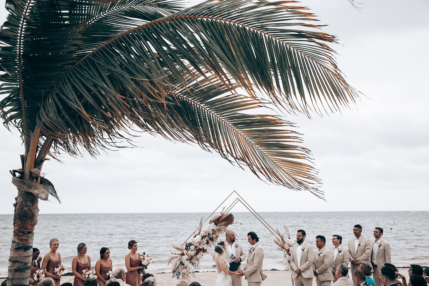 Beach wedding ceremony setup with palm trees and ocean backdrop on a cloudy day.