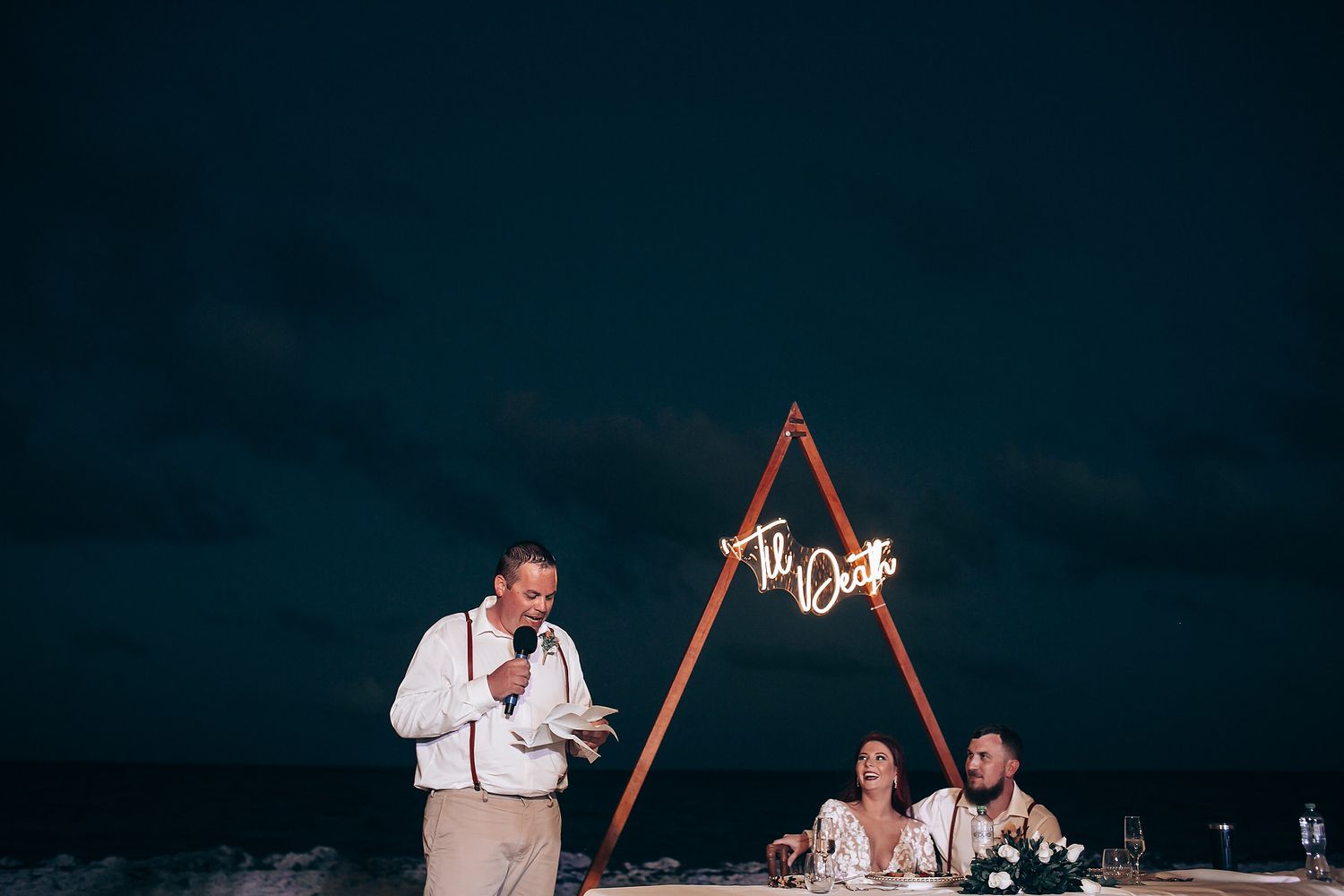 Evening beach reception with wooden arch and string lights creating warm atmosphere.