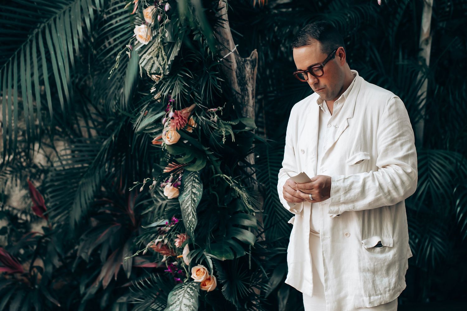 Person in white robe checking phone in tropical garden setting with palm trees and flowers.
